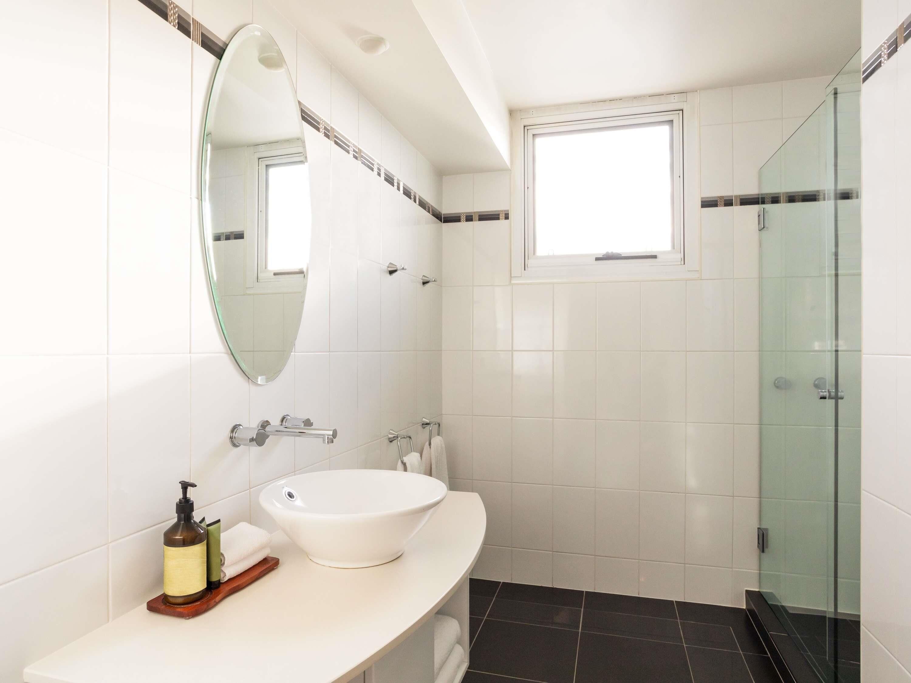A modern bathroom with a white sink, a mirror, a window, and a glass shower enclosure.