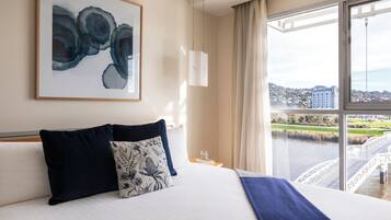 A hotel room with a large bed, a city view through a window, and a framed abstract painting on the wall.