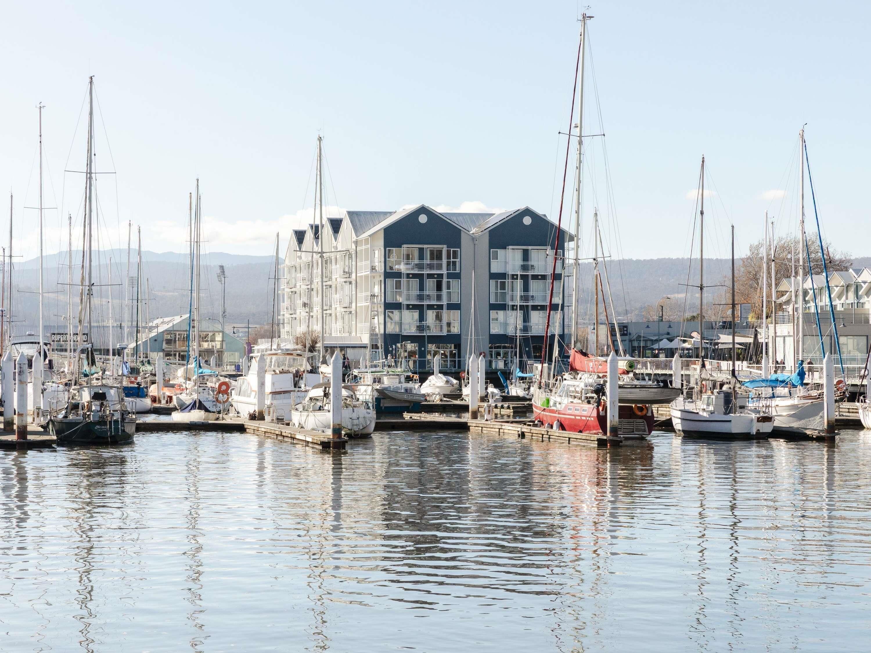 A marina with multiple boats docked, a building with balconies, and a mountainous backdrop.