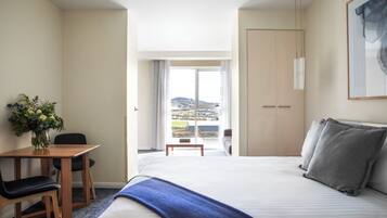 A hotel room with a bed, a desk with a chair, a small table, and a view of a cityscape through a large window.