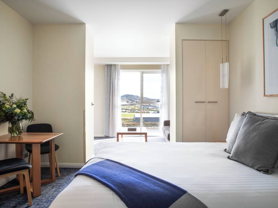 A hotel room with a bed, a desk with a chair, a small table, and a view of a cityscape through a large window.