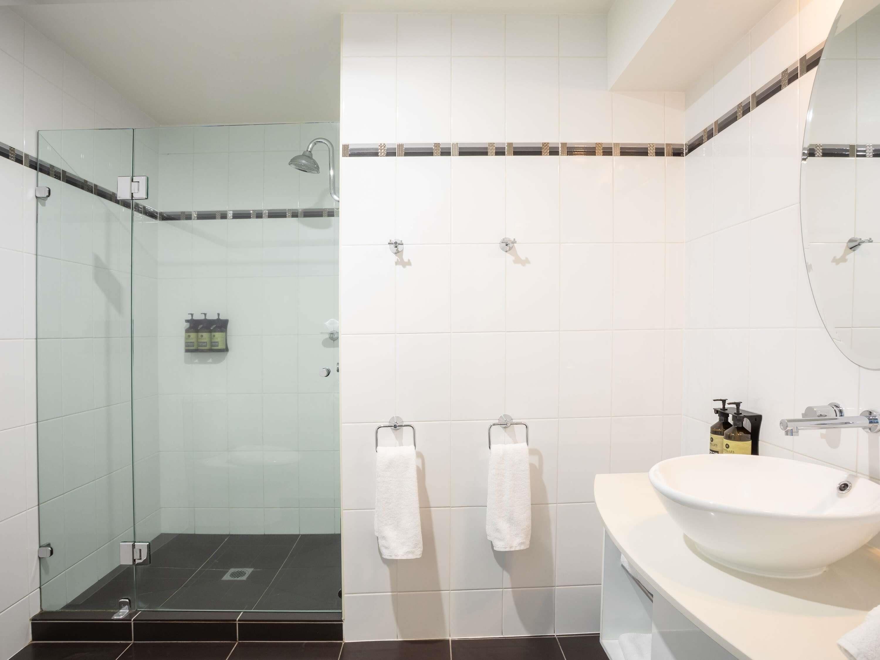 Casa de banho com chuveiro envolto em vidro, azulejos brancos, lavatório branco e duas toalhas penduradas em ganchos.