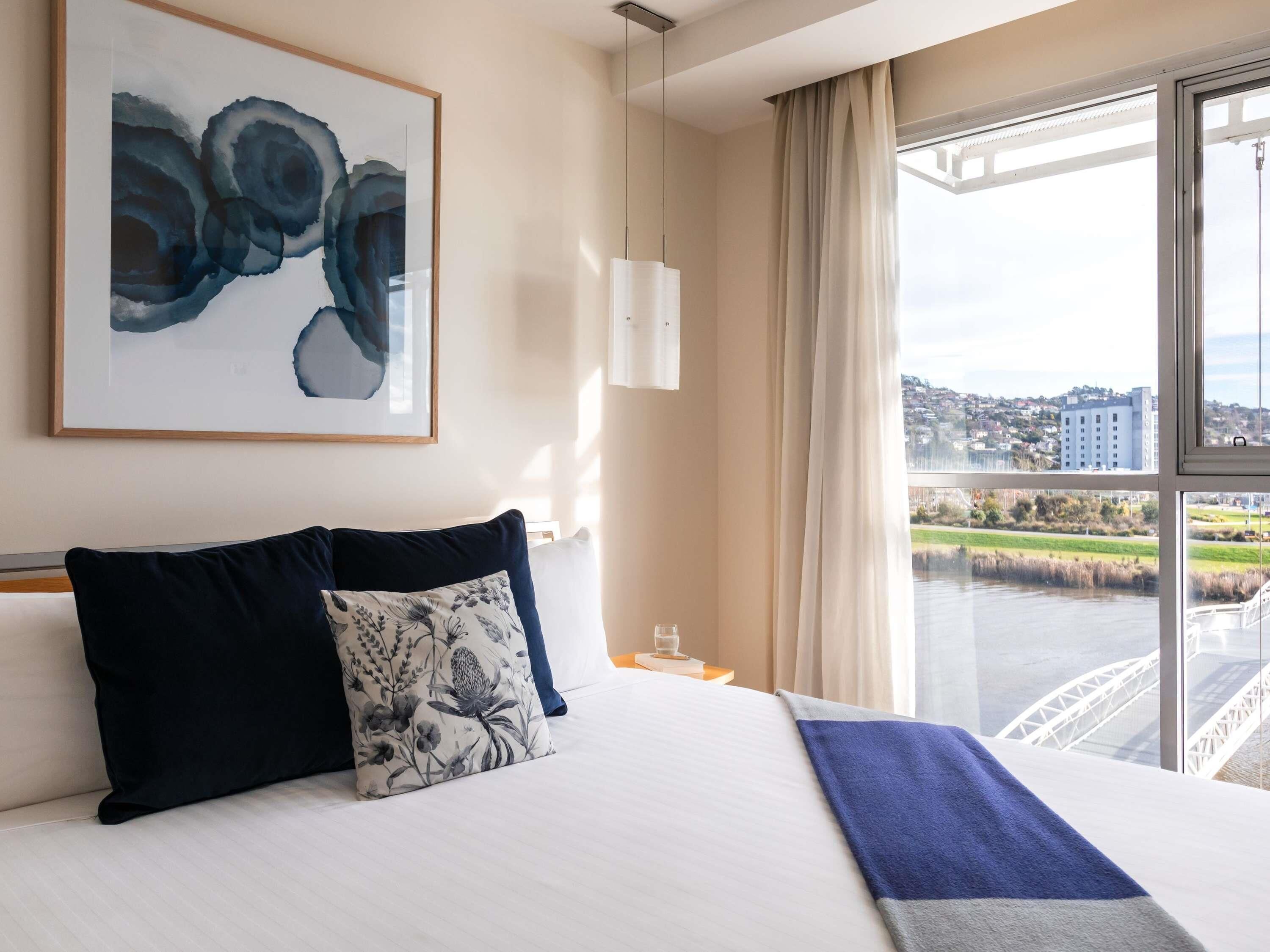 A hotel room with a bed, a large window with a city view, a framed abstract painting, and a nightstand with a glass.