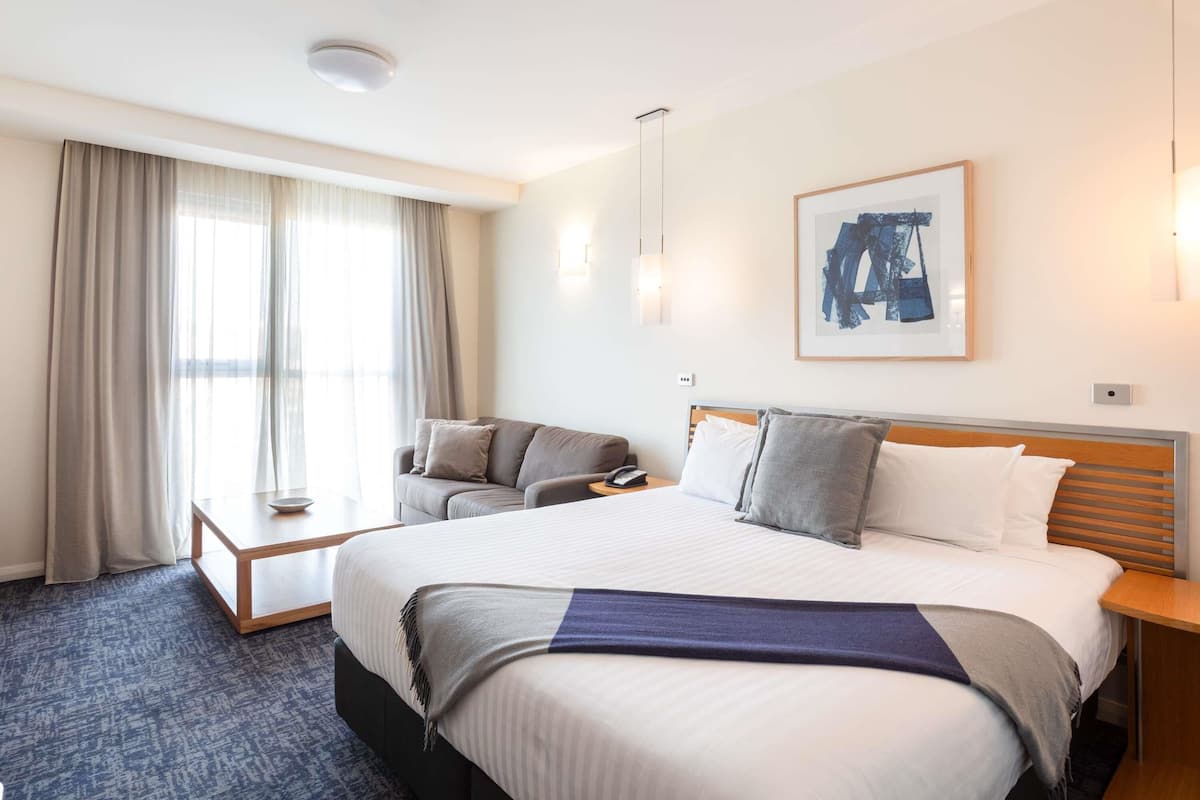 A hotel room with a large bed, a sofa, a bedside table, a lamp, a framed picture on the wall, and a window with curtains.