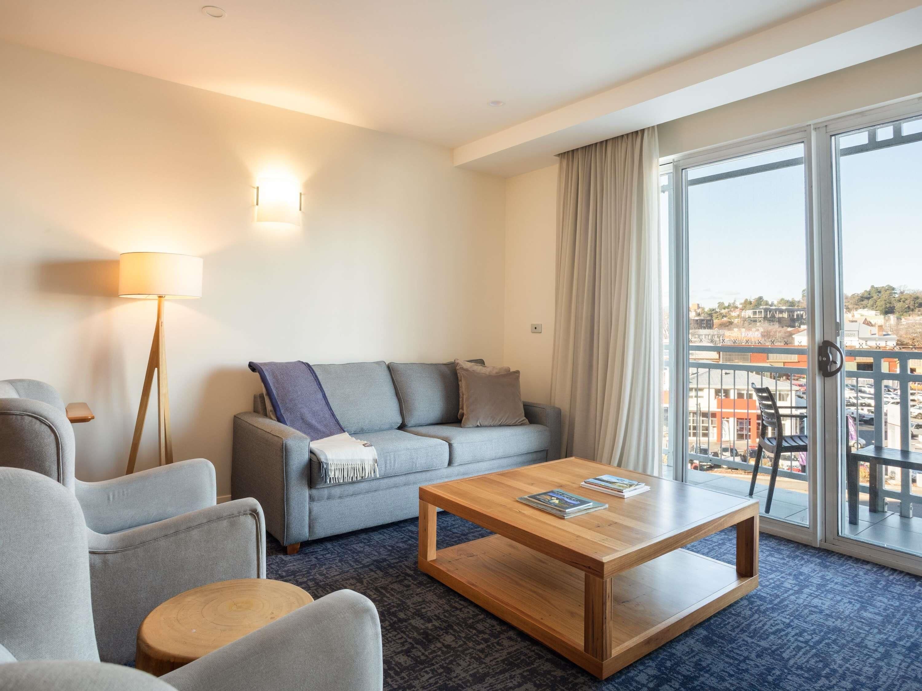 A modern hotel room with a sofa, armchairs, a coffee table, a floor lamp, and a large window with a view of the city.