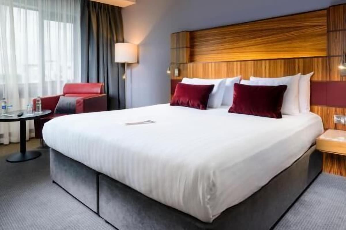 A hotel room with a large bed, a red armchair, a small table, and a lamp.
