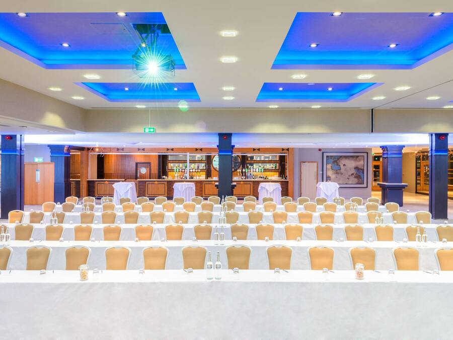 Un salón de conferencias con filas de mesas y sillas, una zona de bar e iluminación azul en el techo.