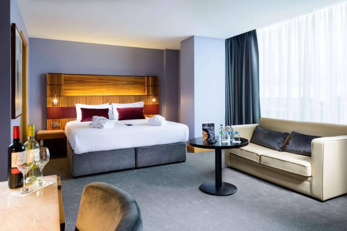 Habitación de hotel con cama, sofá, una pequeña mesa con botellas y vasos, y una ventana con cortinas.