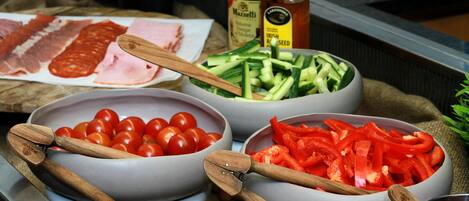 Una encimera de cocina con cuencos de tomates, pepinos y pimientos rojos, una tabla de cortar con carnes en rodajas, y botellas de especias.