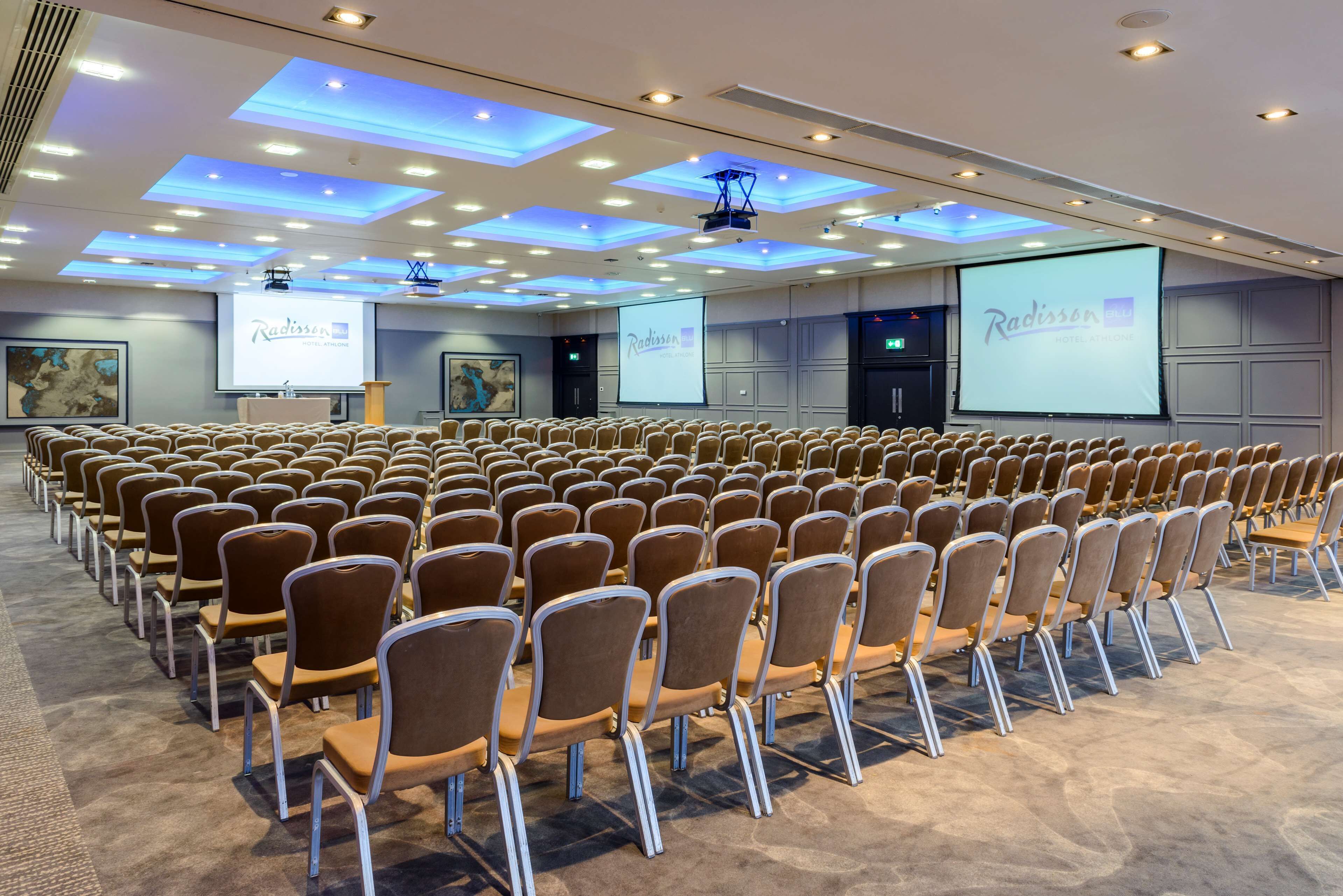 Un salón de conferencias con filas de sillas, un atril y una pantalla grande mostrando el logotipo del Hotel Radisson.