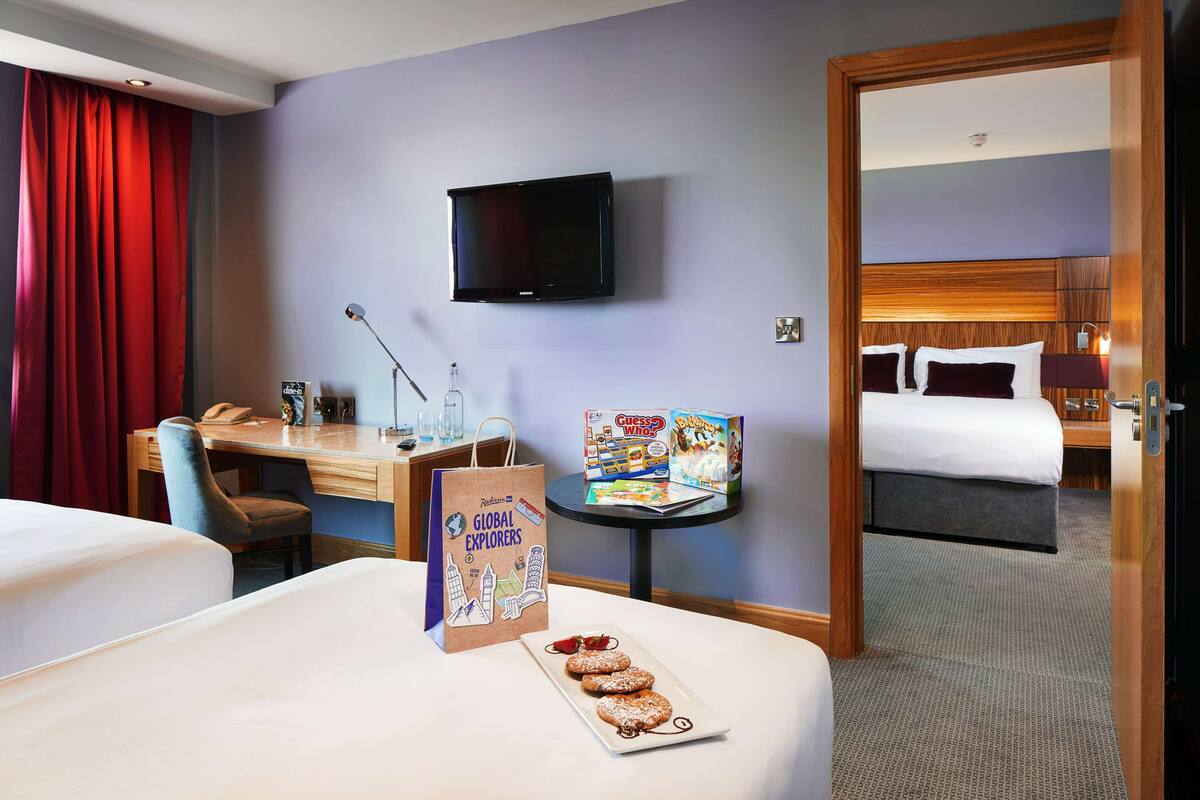 Habitación de hotel con cama, escritorio, silla, televisor, una mesita con un juego de mesa y una puerta que da a otra habitación.