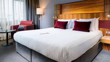 A hotel room with a large bed, a red armchair, a small table, and a lamp.