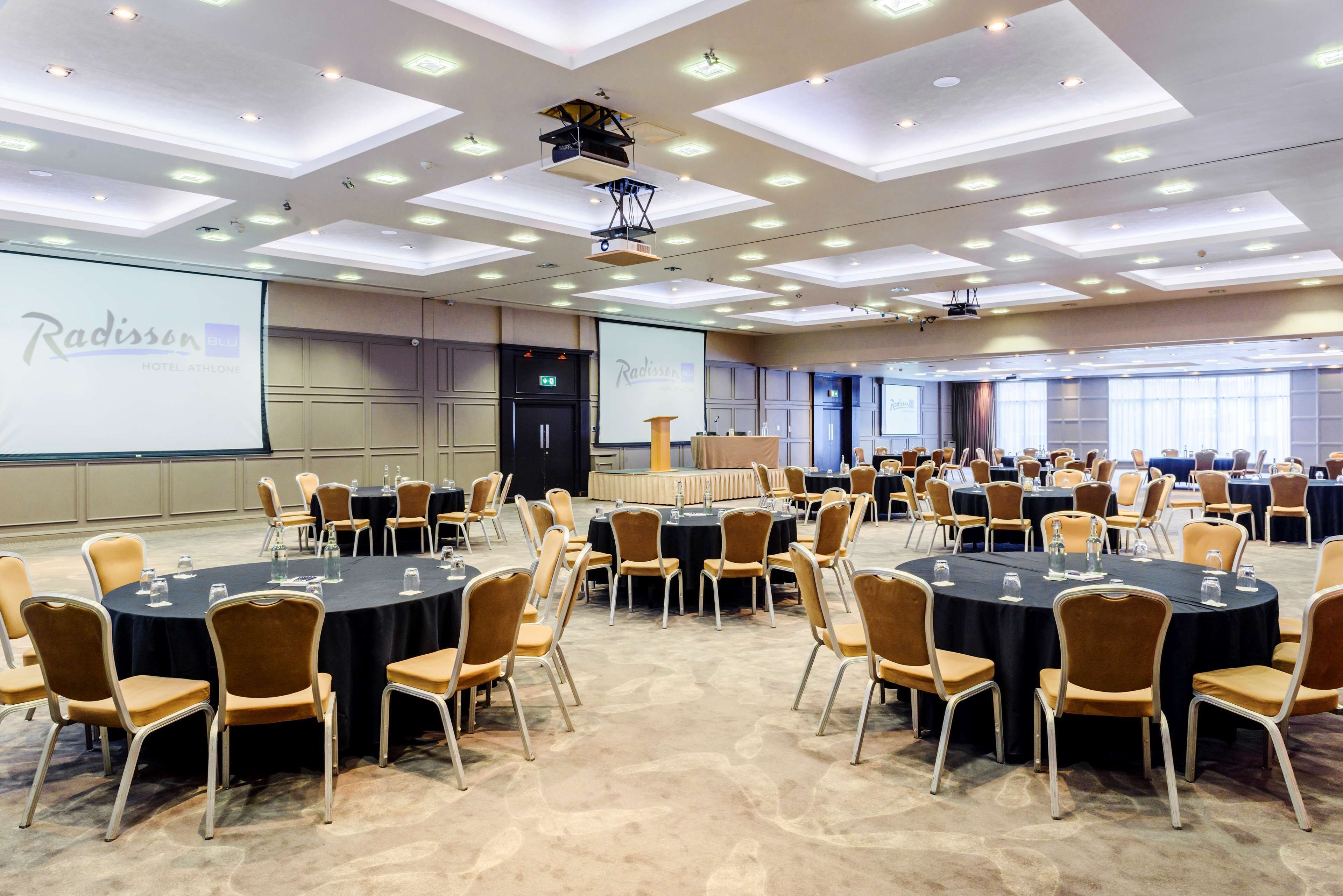 A conference room with multiple round tables and chairs, a projector screen, and a stage area.