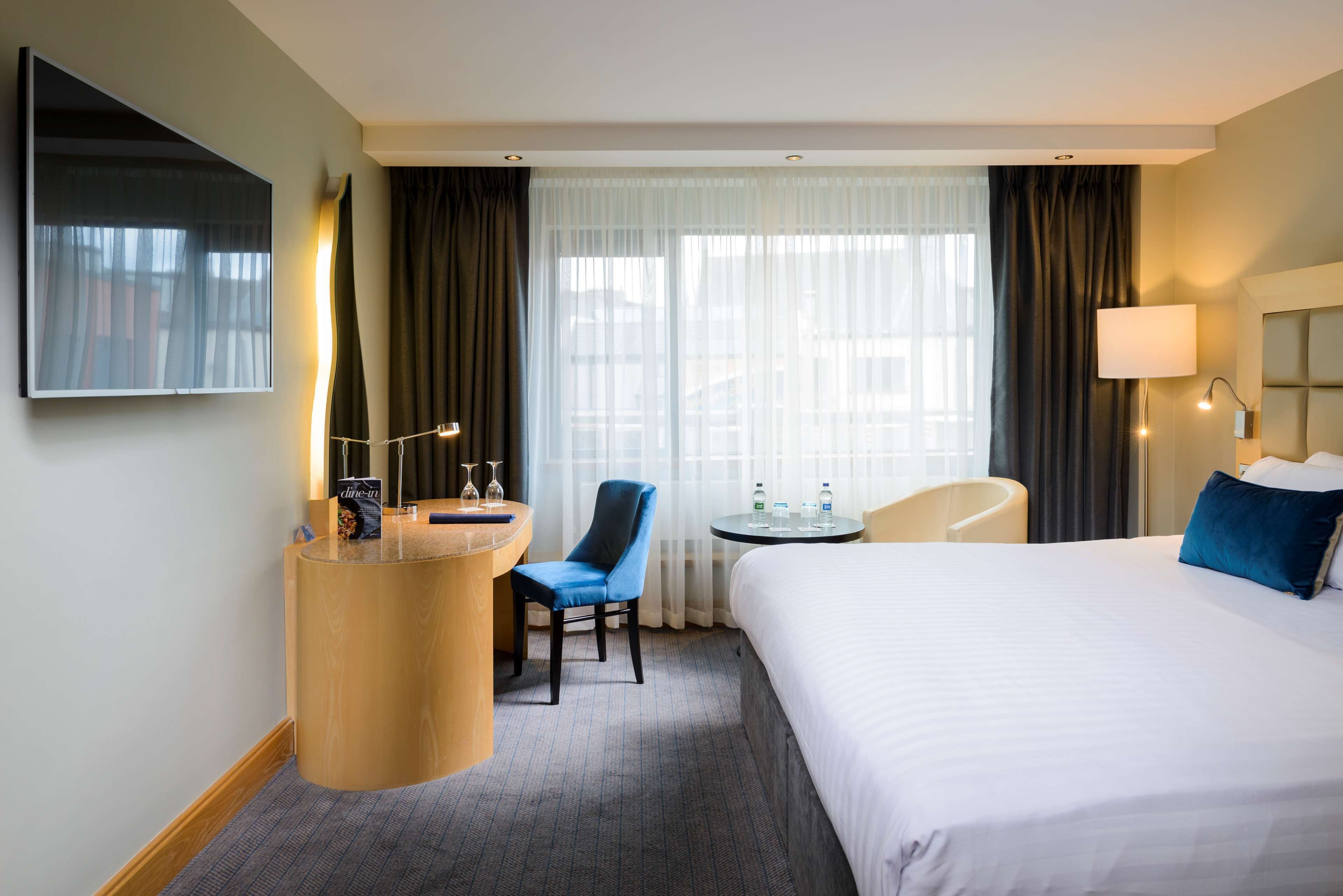A hotel room with a bed, a desk with a chair, a small table, a TV mounted on the wall, and a window with curtains.
