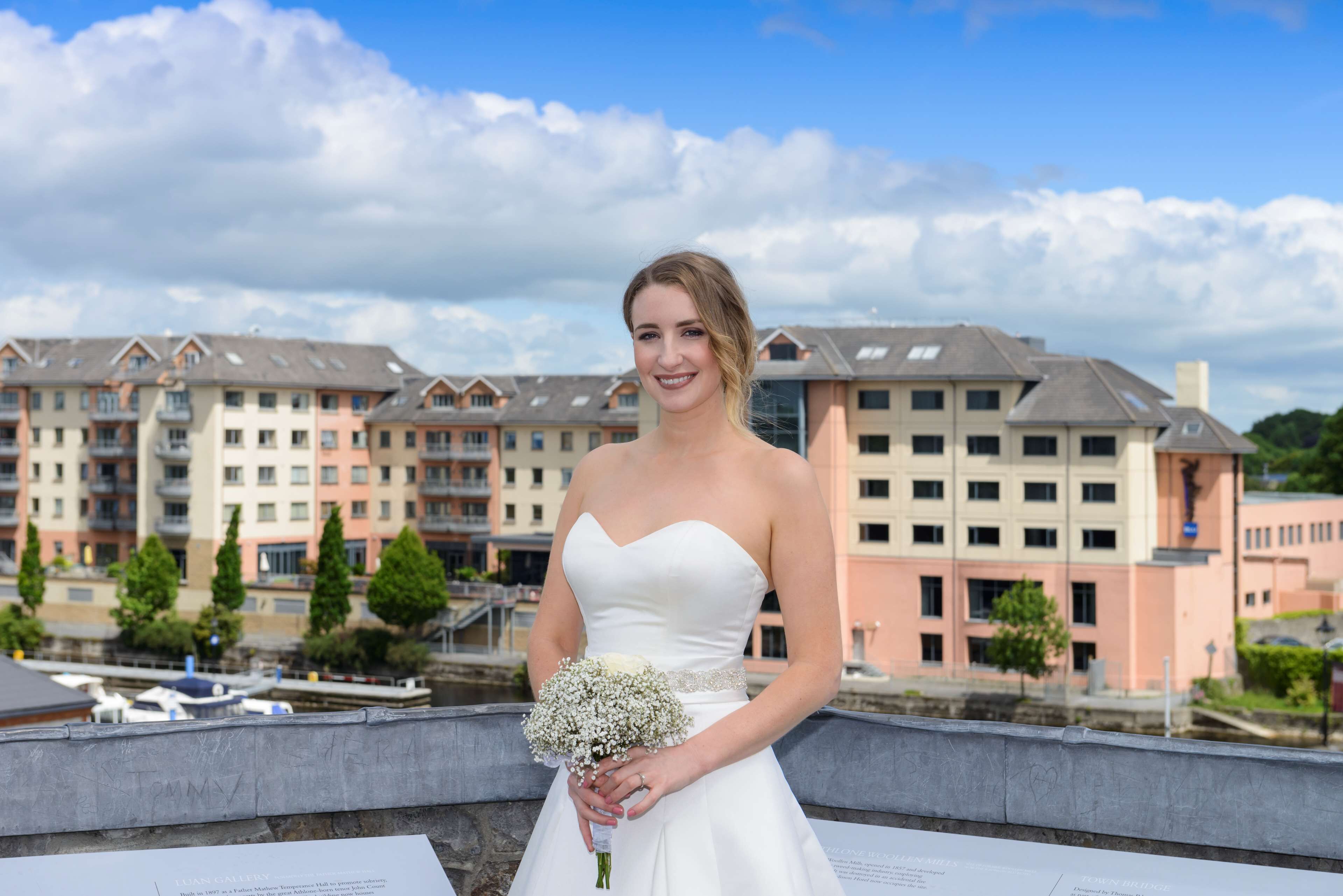 Eine Braut in einem weißen Brautkleid hält einen Blumenstrauß, im Hintergrund ist ein Hotelgebäude zu sehen.