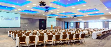 A conference room with rows of tables and chairs, a projector screen, and a Radisson BLU hotel sign.
