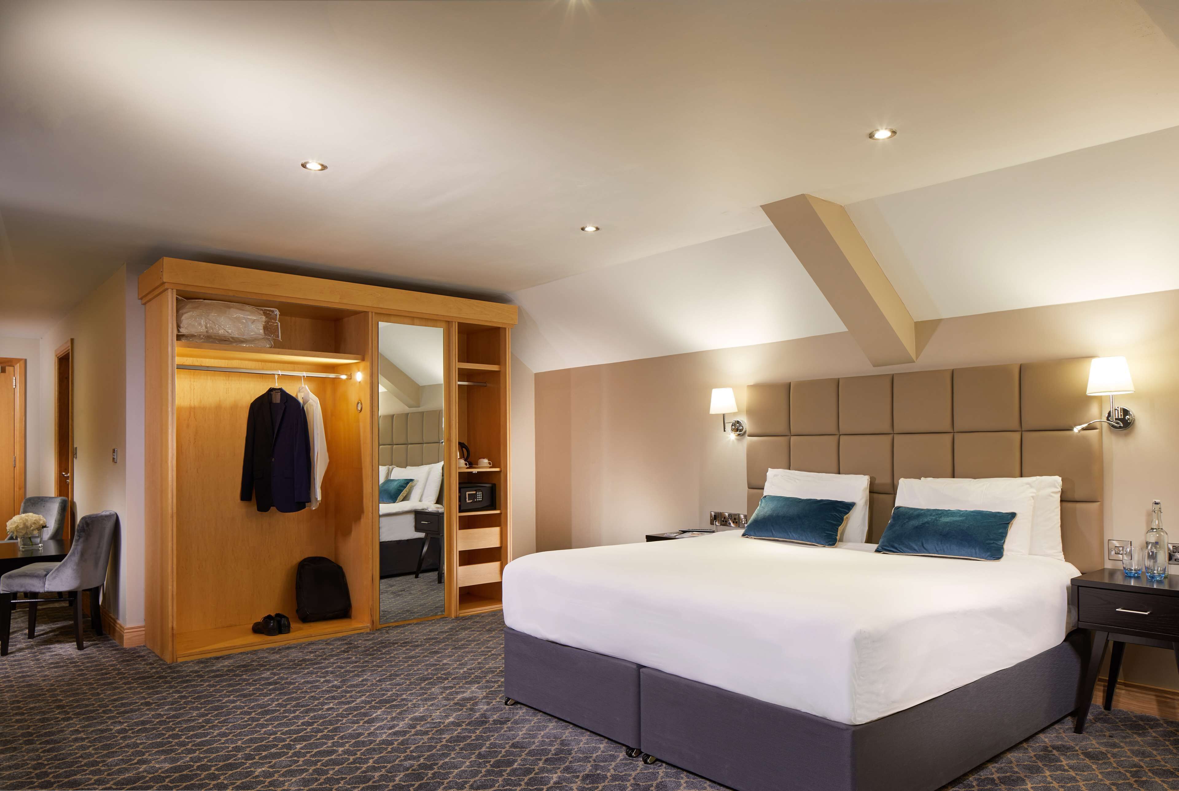 A hotel room with a large bed, a walk-in closet, a desk with a chair, and a small table with a vase.