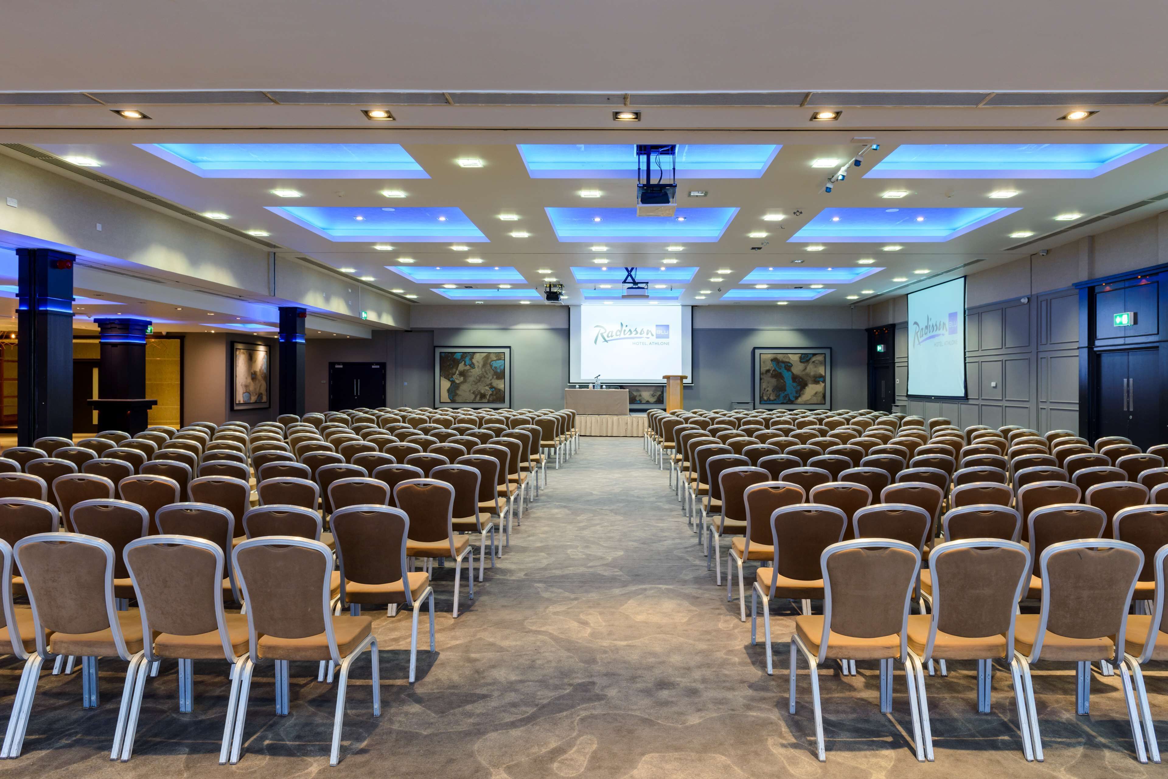 Une salle de conférence avec des rangées de chaises disposées face à un écran de présentation, une scène avec un pupitre et un plafond doté d’un éclairage encastré.
