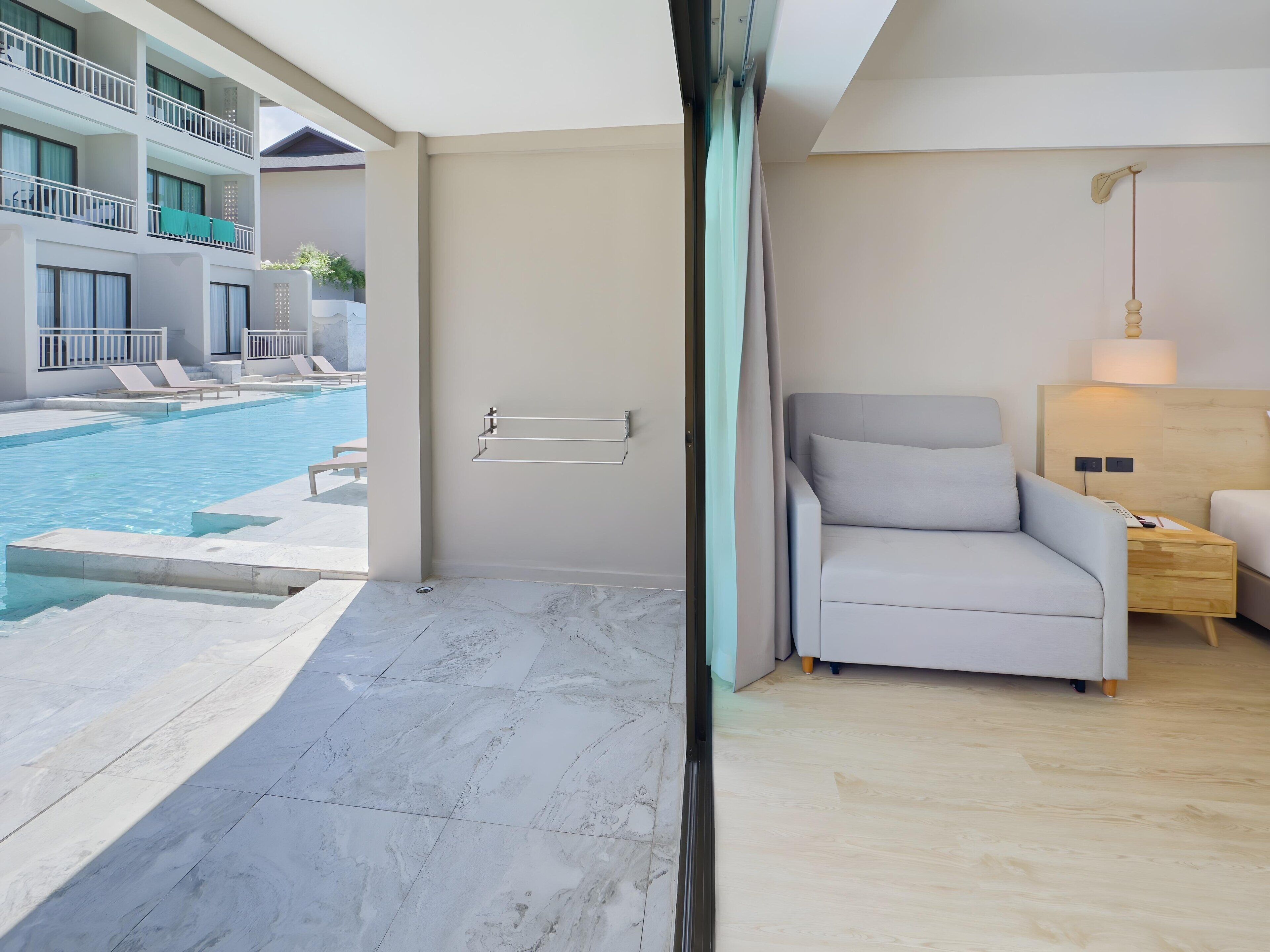 A modern hotel room with a view of a swimming pool, a grey sofa, a wooden nightstand, and a wall-mounted towel rack.
