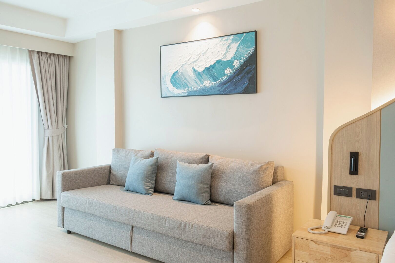 A modern hotel room with a grey sofa, a painting of a wave, a wooden nightstand with a phone, and a window with curtains.