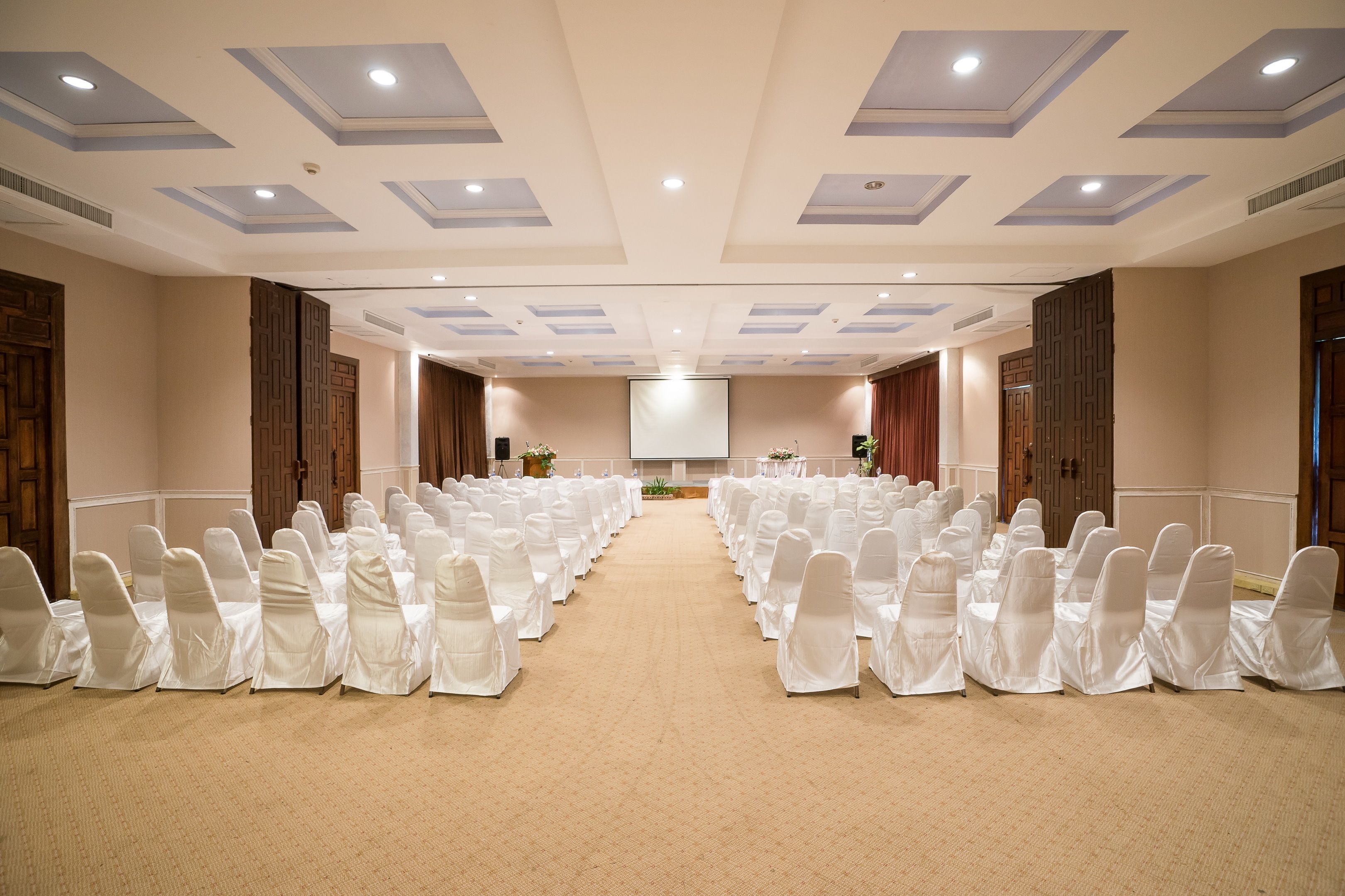 A conference room with rows of white-covered chairs, a projector screen, and a carpeted floor.