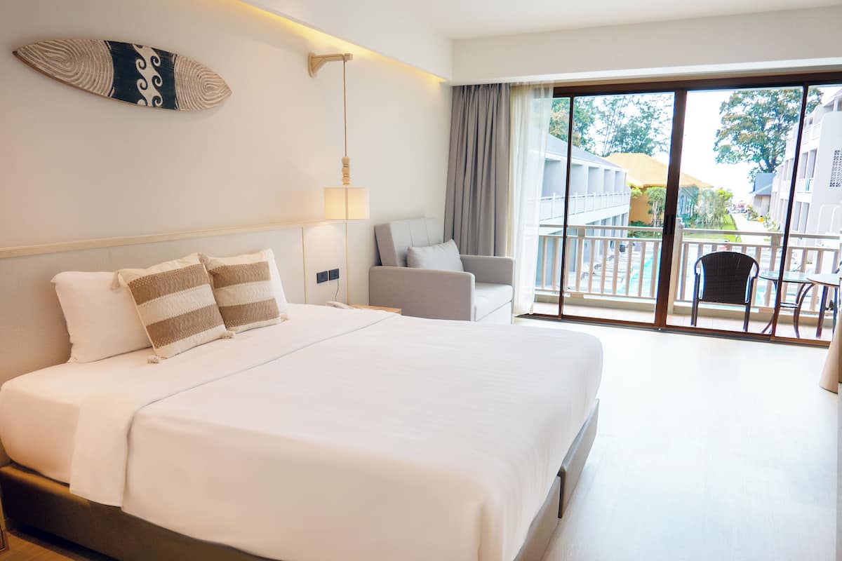 Habitación de hotel con una cama grande, una mesita de noche, una lámpara, una silla y un balcón con vistas a edificios y árboles.