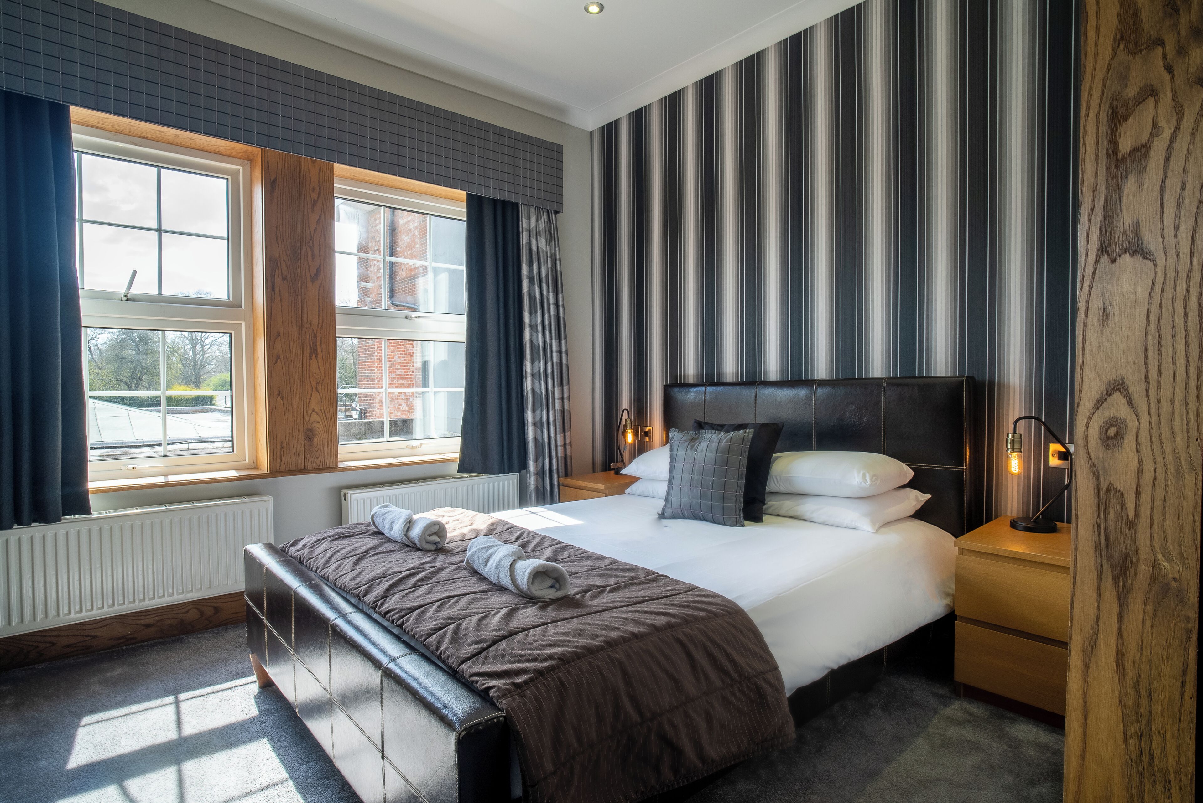 A modern hotel room with a large bed, a wooden headboard, a nightstand, and a window with curtains.