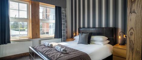 A modern hotel room with a large bed, a wooden headboard, a nightstand, and a window with curtains.