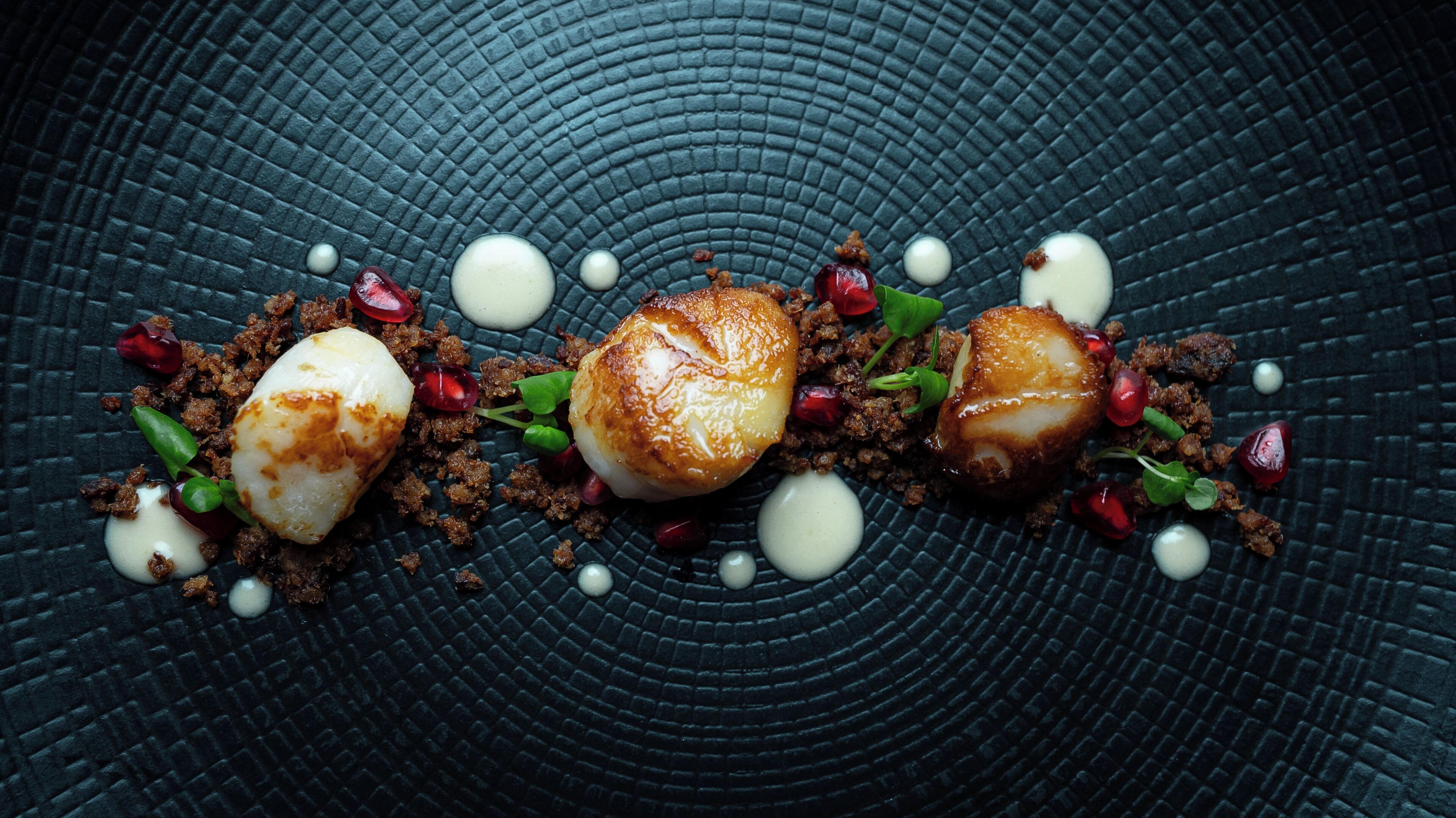 Une assiette contenant trois noix de Saint-Jacques, décorée de graines de grenade et de jeunes pousses, le tout présenté sur une assiette sombre à la texture particulière.