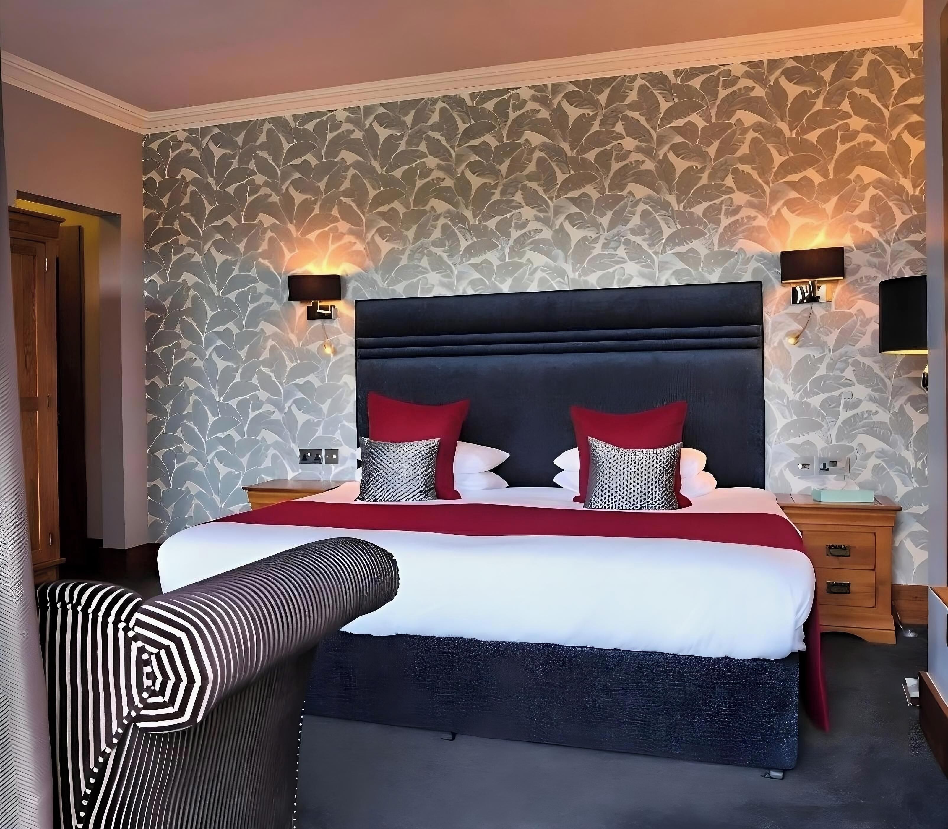 A neatly made bed with a black headboard, red and white pillows, and a wooden bedside table with a lamp.