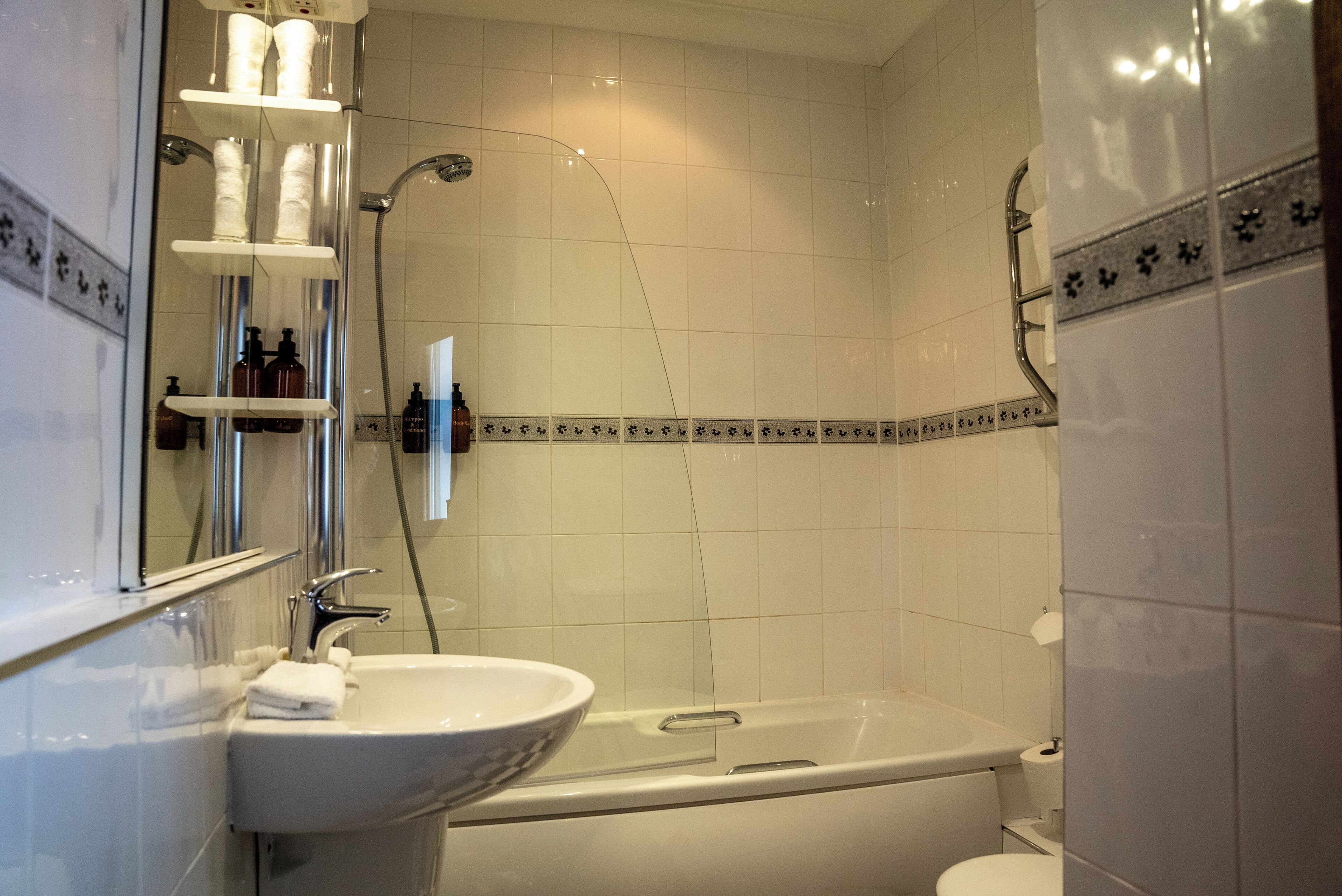 Un baño con una bañera, un lavabo y un inodoro. Hay estantes con artículos de aseo y un espejo sobre el lavabo.