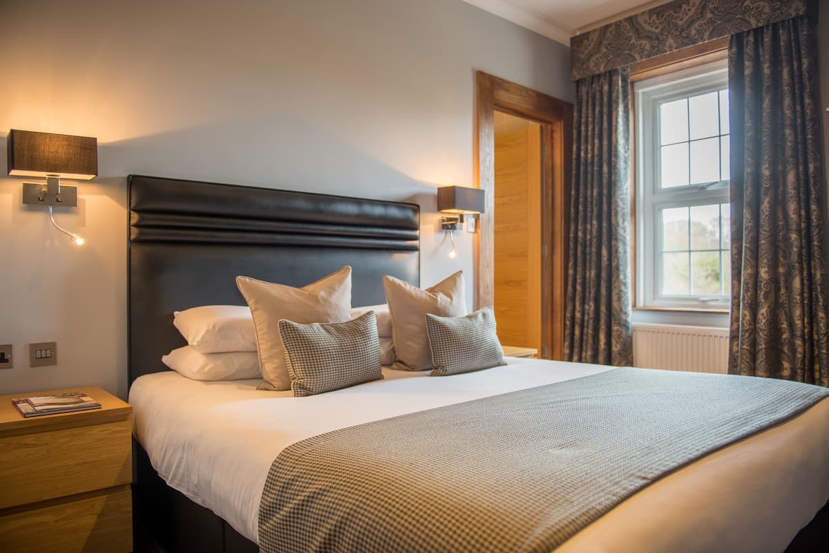 A neatly made bed with a headboard, two bedside lamps, a nightstand, and a window with curtains.