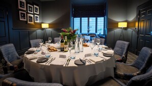 Mesa redonda para seis personas, dispuesta con mantel blanco, rodeada por seis sillas tapizadas. Hay copas de vino, vasos de agua y un centro de mesa con flores. La habitación tiene una decoración en tonos oscuros, con paredes azules y una ventana con vistas.