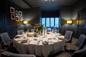 A round dining table set for six with a white tablecloth, surrounded by six upholstered chairs. There are wine glasses, water glasses, and a centerpiece with flowers. The room has a dark color scheme with blue walls and a window with a view. - Balmer Lawn Hotel (Brockenhurst)