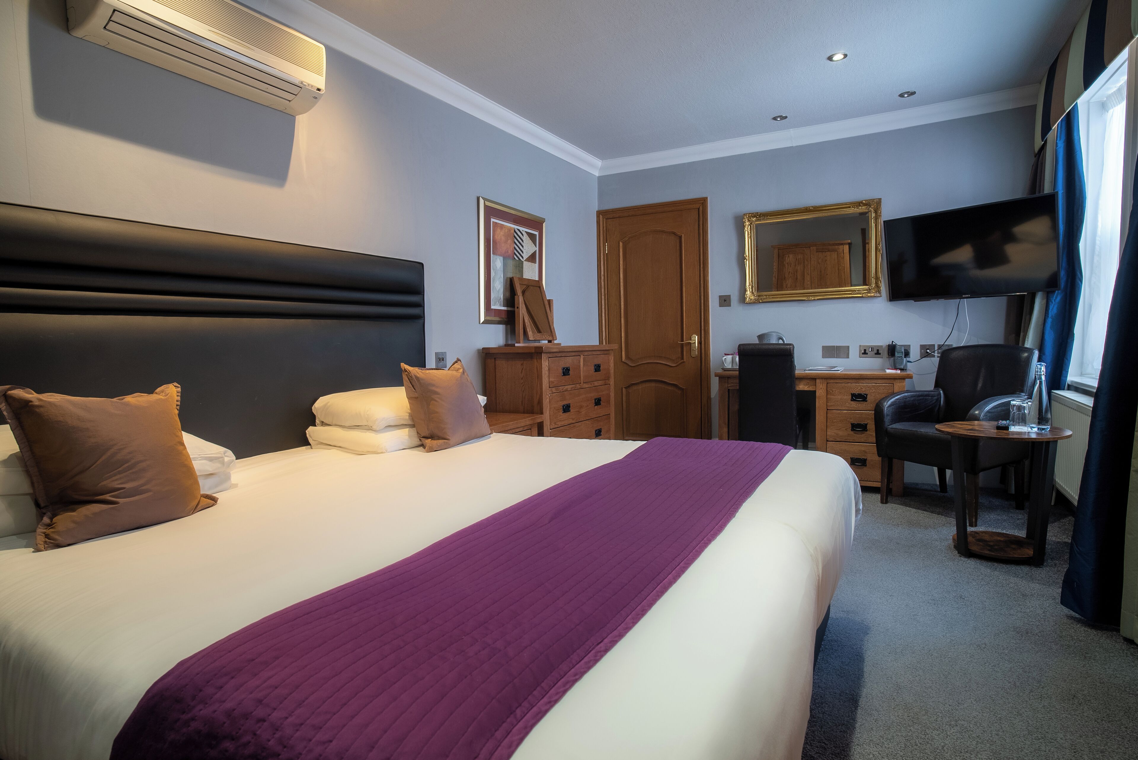 A hotel room with a large bed, a desk, a chair, a television, and a wardrobe.