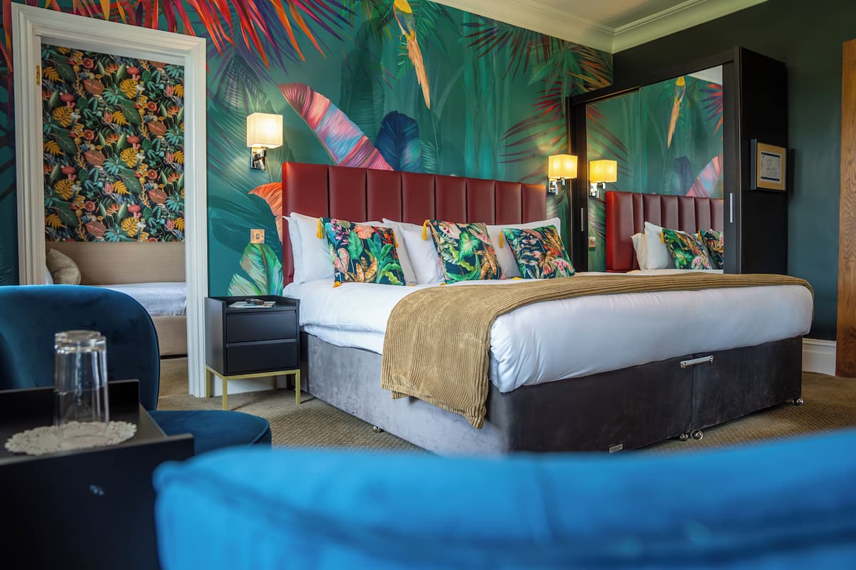 A hotel room with a large bed, a nightstand, a blue armchair, and a tropical-themed wall mural.