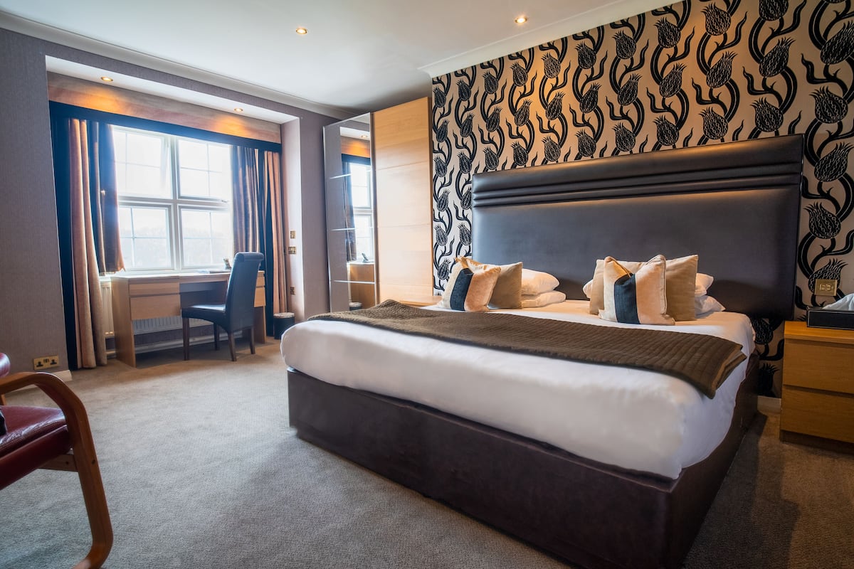 A hotel room with a large bed, a desk, and a chair. There is a window with curtains, a patterned wall, and a nightstand.