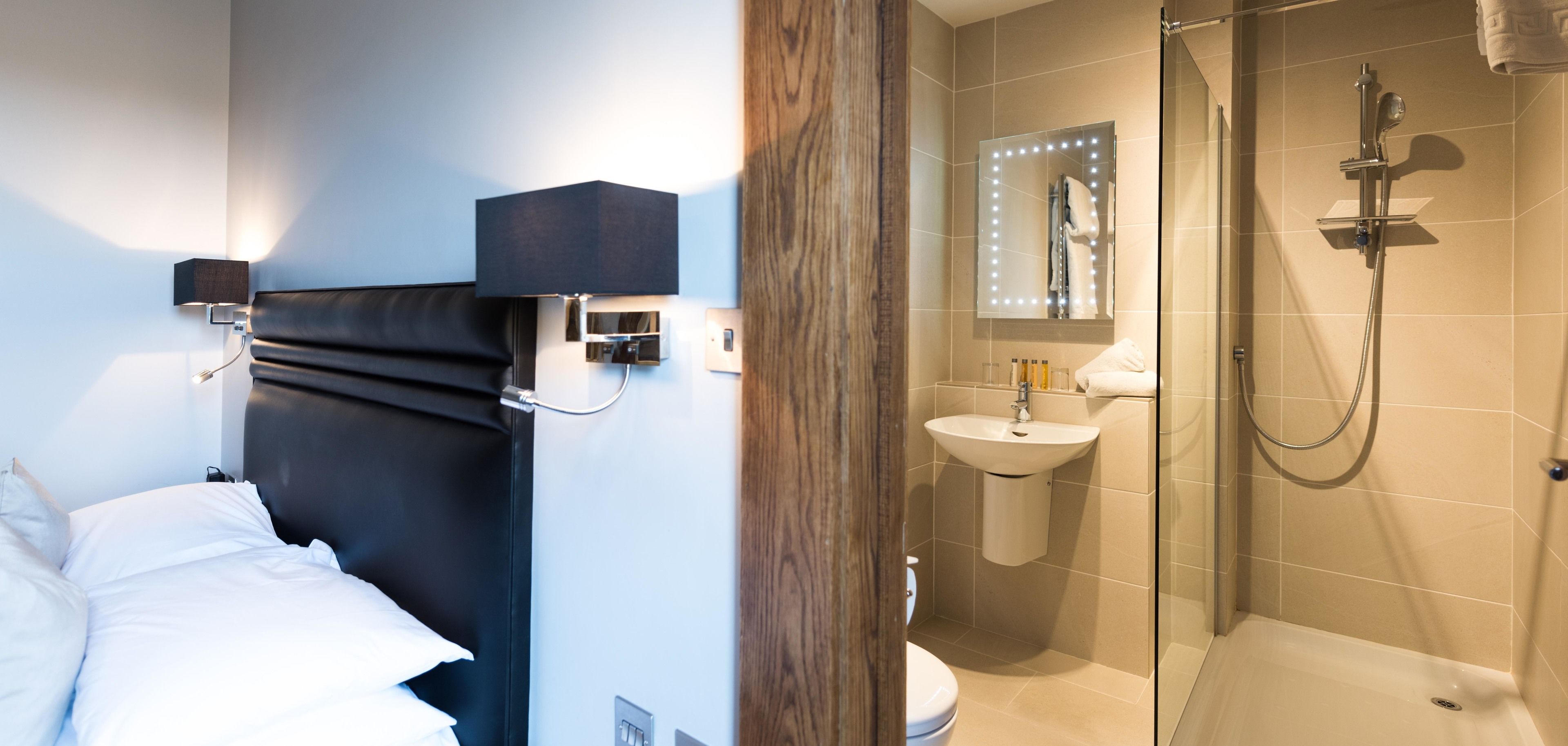 A modern hotel room with a bed, bedside lamp, and a bathroom with a shower, sink, and mirror.