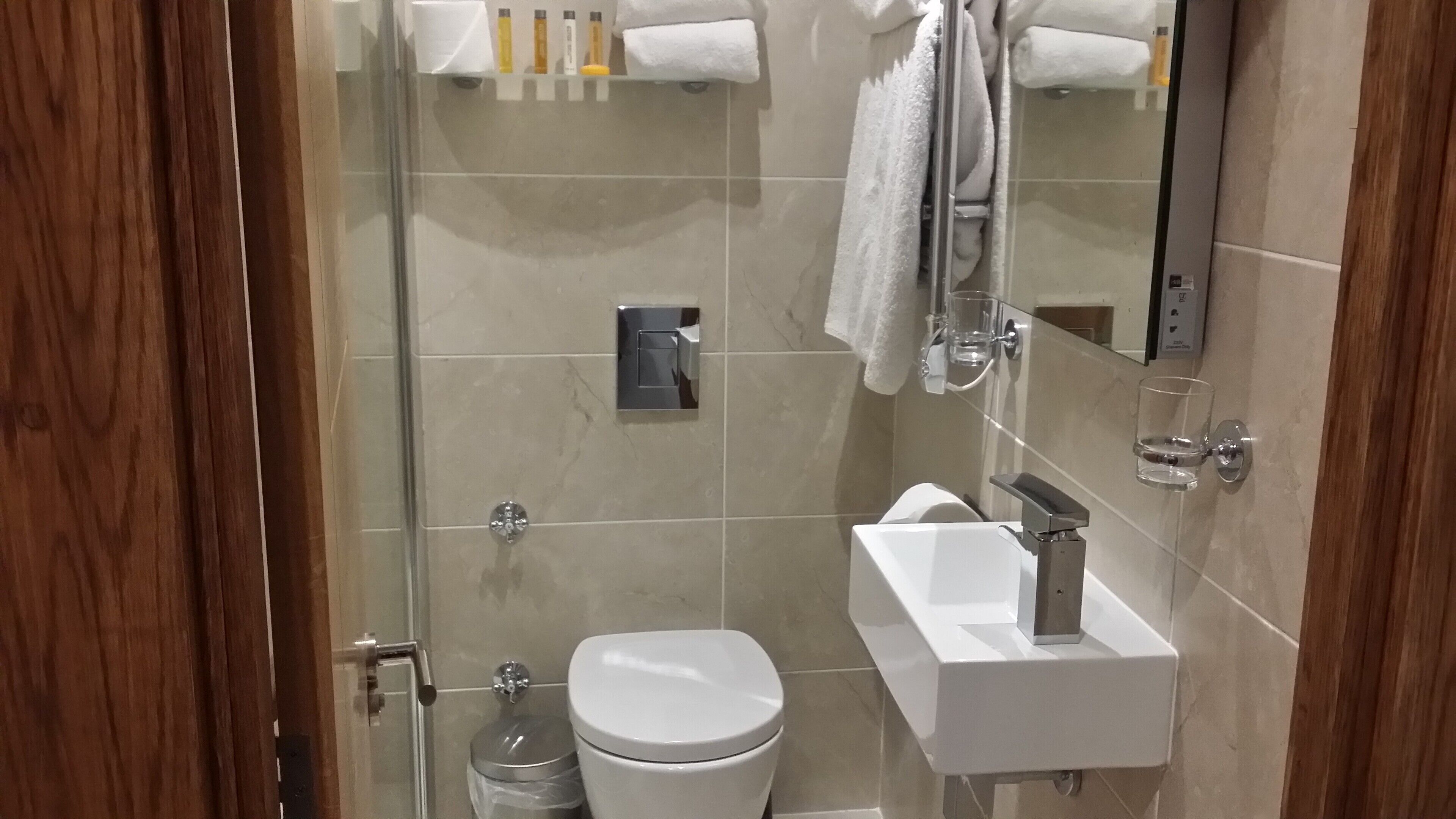 A modern bathroom with a glass shower enclosure, a white toilet, a sink with a mirror, and toiletries on a shelf.