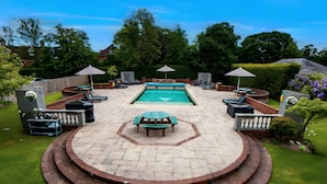 Un patio circular con piscina, rodeado de sillones de descanso y sombrillas, en un exuberante jardín.