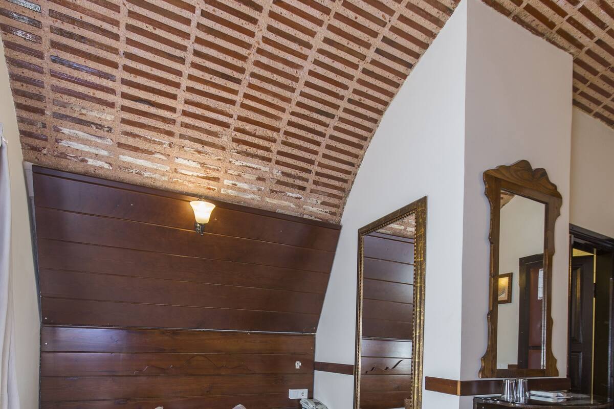 A bedroom with a wooden headboard, a large bed with a patterned bedspread, a nightstand, a mirror, and a bench.