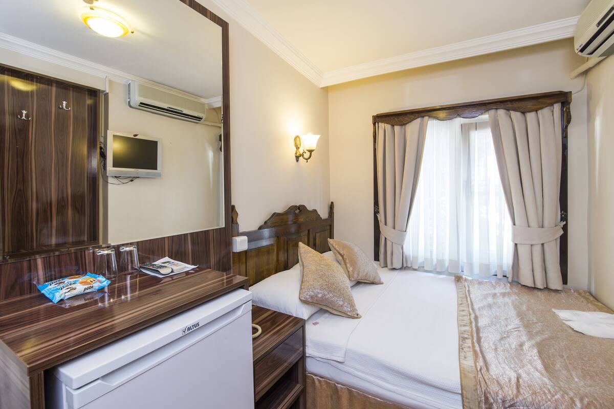 A hotel room with a bed, a desk with a computer, a mirror, an air conditioner, and a window with curtains.