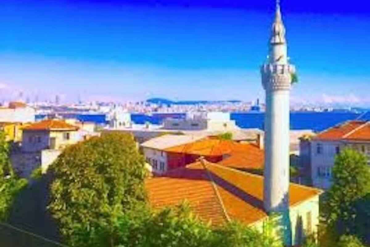 A coastal cityscape with a prominent white tower, likely a mosque, surrounded by residential buildings with red-tiled roofs.