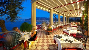 An outdoor dining area with tables and chairs, a checkered floor, and a view of the sea and mountains in the background. - Hotel Minerva (Sorrento)