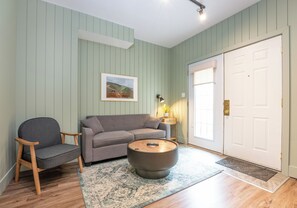 Une pièce avec un canapé gris, une chaise en bois, une table basse ronde et une porte avec un panneau blanc.