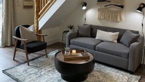 Een moderne woonkamer met een grijze bank, een ronde salontafel, een houten stoel, een trap en een wandlamp.