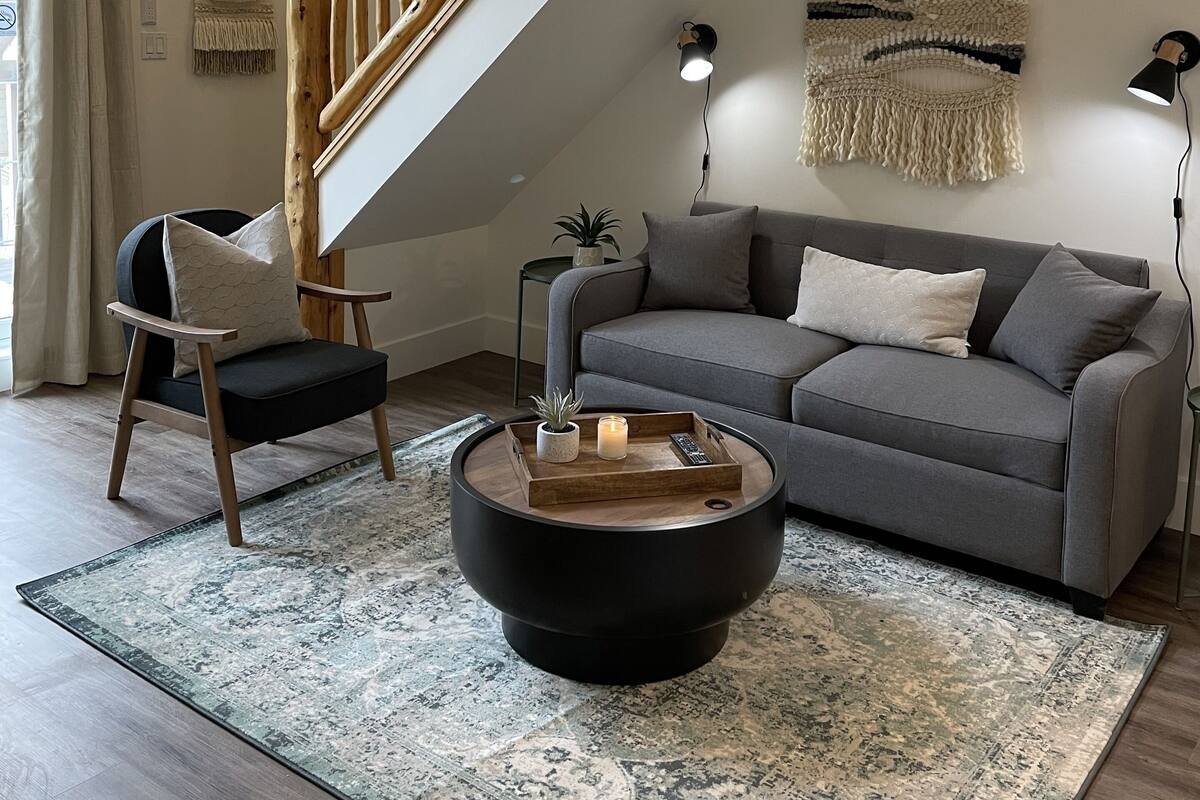 Un salon moderne avec un canapé gris, une table basse ronde, une chaise en bois, un escalier et une lampe fixée au mur.