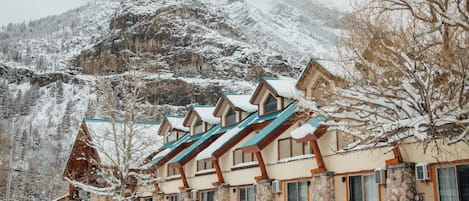 A row of chalet-style buildings with stone walls and wooden roofs, set against a snowy mountain backdrop.