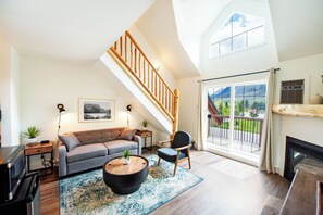 Un salon spacieux avec un plafond haut, un escalier en bois, un canapé, une table basse et une vue dégagée sur l’extérieur grâce à de grandes fenêtres.