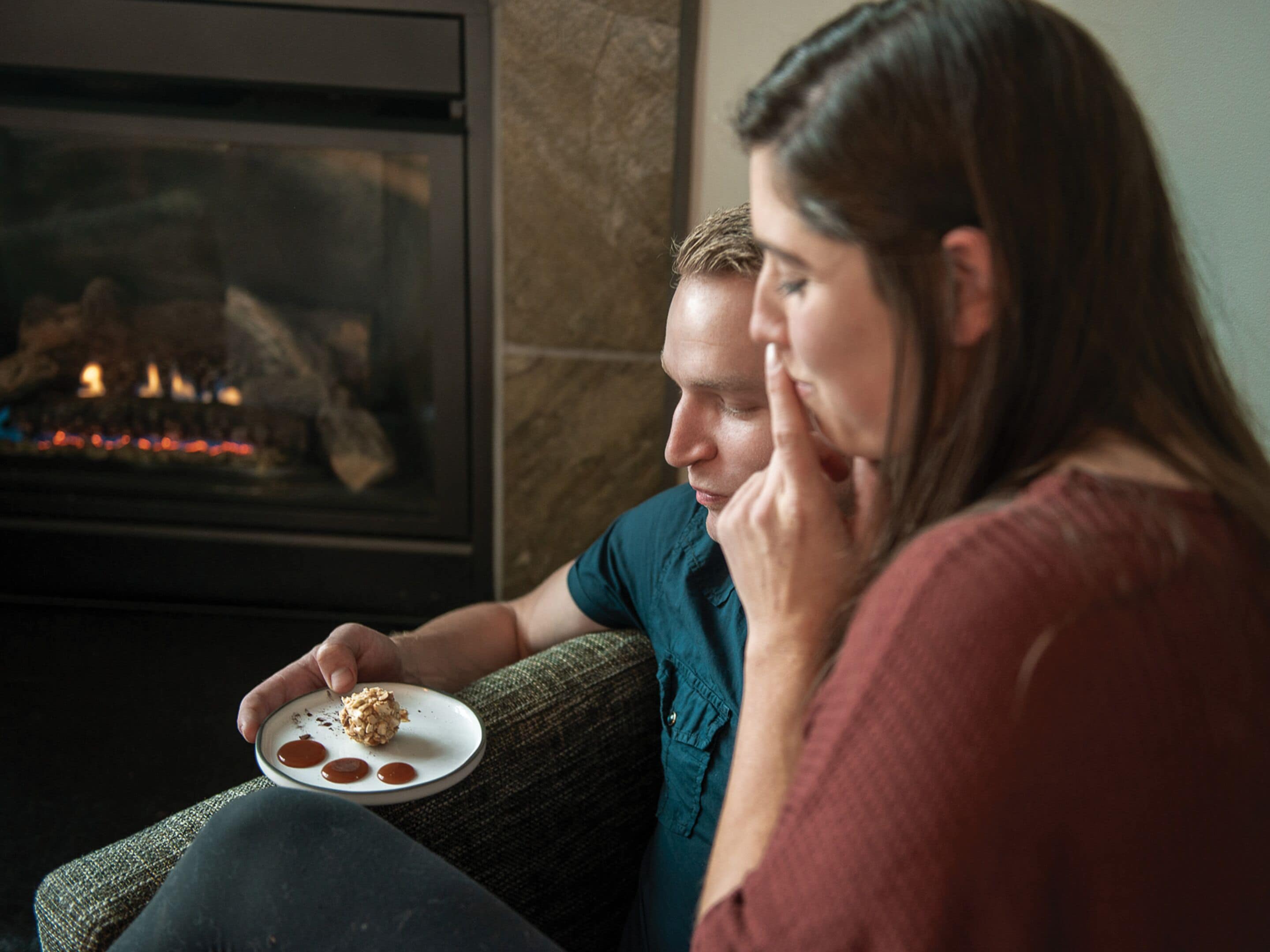 A person holding a plate with food, sitting on a couch in a room with a fireplace.